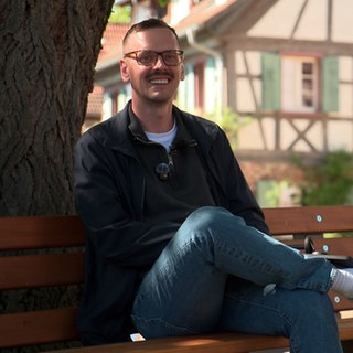 Patrik Lehmann sitzt auf einer Parkbank in einem Dorf, die im Schatten eines Baumes steht. Er lächelt in die Kamera.