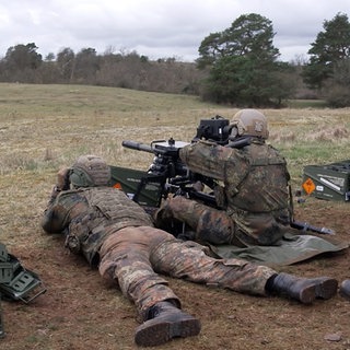 Mehrere Soldaten in Tarnanzügen sitzen auf einer Wiese. Einer von ihnen sitzt an einer großen Haubitze, ein anderer liegt links neben ihm. Rechts von ihnen sitzt ein Soldat mit einem Fernglas auf einem Stuhl.