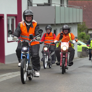 Sechs Menschen fahren auf Mofas eine Straße hoch. 