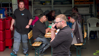 Ein Mann im Vordergrund trägt Arbeitskleidung und telefoniert. Im Hintergrund sitzen mehrere Menschen auf Bierbänken und essen.