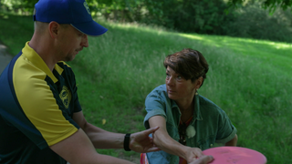 Sonja Faber-Schrecklein hat eine Frisbee in der Hand. Ein Mann hält sie am Arm, um ihr zu zeigen, wie man die Frisbee richtig wirft. 
