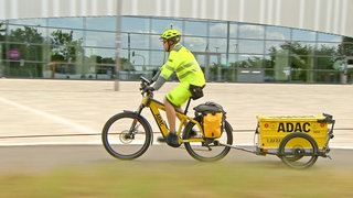Heiko Graftl sitzt auf einem gelben Fahrrad mit einem Anhänger. Auf dem Anhänger steht ADAC. Er trägt eine Schutzweste und einen Helm und fährt nach links.