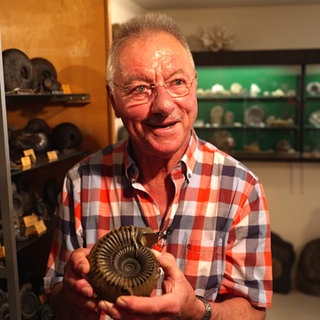 Elmar Scherer steht in einem dunklen Raum mit verschiedenen Fossilien. In seinen Händen hält er einen großen Ammoniten und hält ihn in die Kamera. Er lächelt dabei begeistert.