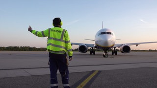 Simon Fauth steht mit einer neongelben Weste mit der Aufrschrift "Dispatcher" vor einem Flugzeug und zeigt mit dem linken Daumen nach oben.