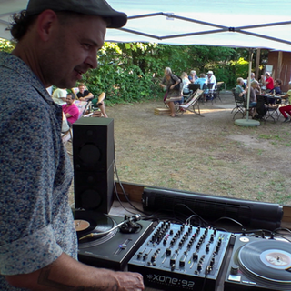 Ein Mann steht an einem DJ-Pult. Im Hintergrund sitzen Menschen in einem Biergarten.