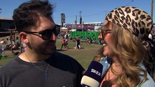 Jannik und Patricia sind auf dem Glückgefühle Festival auf dem Hockenheimring. Sie tragen Sonnenbrillen und schauen sich an.