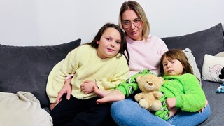 Eine Mutter sitzt mit ihren zwei Töchtern und dem Teddy Bäri auf der Couch.