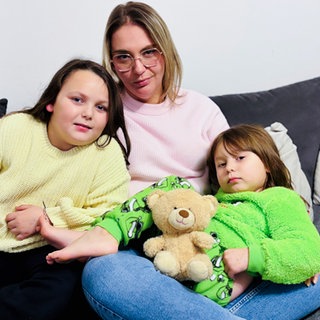 Eine Mutter sitzt mit ihren zwei Töchtern und dem Teddy Bäri auf der Couch.