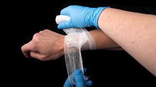 Zwei behandschuhte Hände wickeln eine Mullbinde um ein Handgelenk