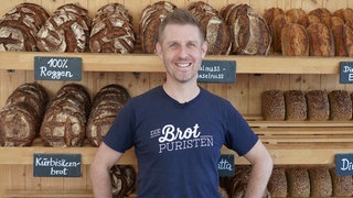 Sebastian Däuwel trägt ein T-Shirt mit der Aufschrift „Die Brotpuristen“ und steht vor einem Brotregal in seiner Bäckerei.