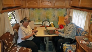 Anka (l.) und Thomas Blödorn arbeiten am Handy und am Laptop in ihrem Wohnwagen