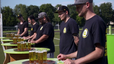 Junge Männer der Football-Mannschaft "Bulldogs" stehen vor Stehtischen mit vollen Biergläsern. Qualität, Geschmack, Nachhaltigkeit von Rothaus Tannenzäpfle
